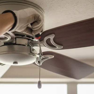 A close-up of a person's hand tightening a screw on a ceiling fan blade bracket with a screwdriver, emphasizing DIY repair.
